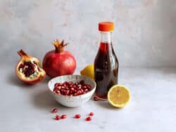 Bottle of homemade grenadine surrounded by a bowl of pomegranate seeds and half a lemon