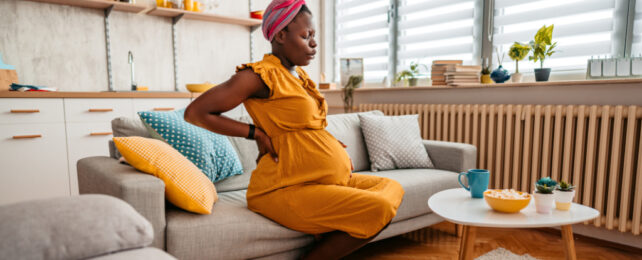 Pregnant woman sitting on couch, holding stomach and back while breathing out.